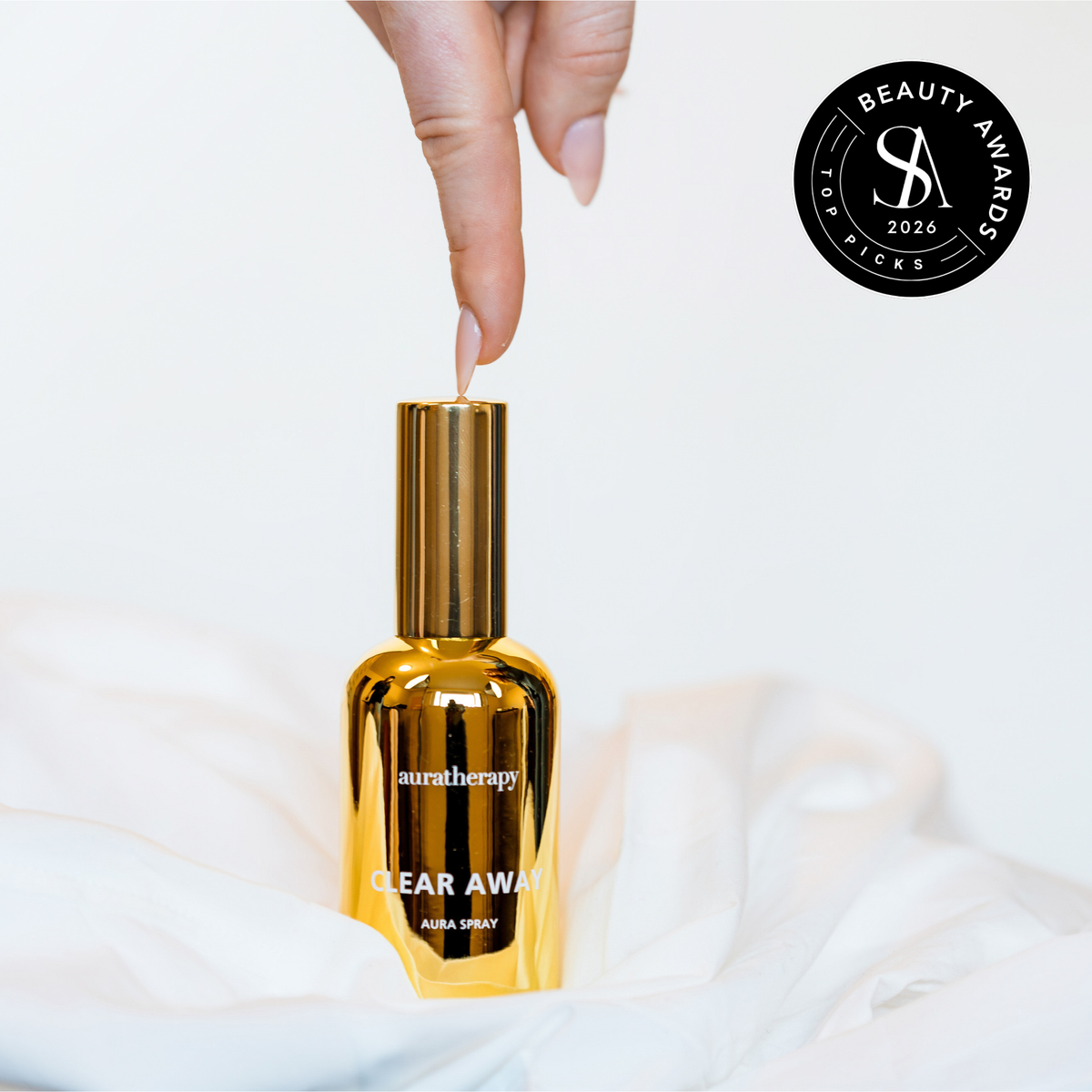 Hand pressing the gold Auratherapy Clear Away Aura Spray bottle on white linens, with the Skin Anarchy 2026 Top Picks Beauty Awards seal