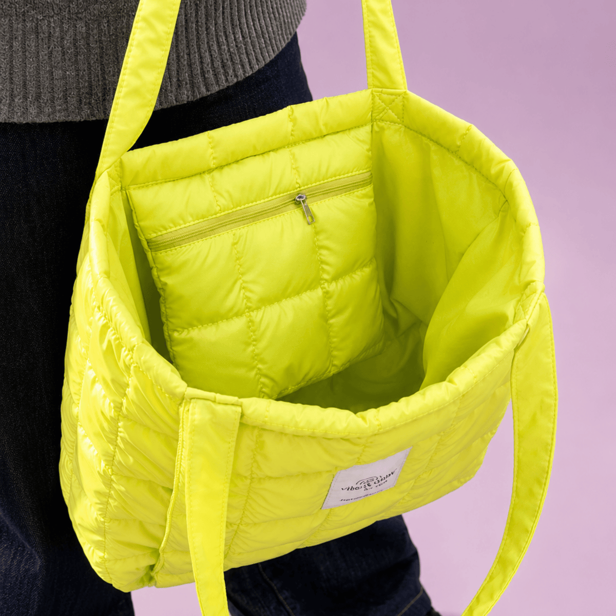 A bright neon yellow quilted puffer tote bag with a matching inner pocket and shoulder straps, shown being worn by a person in a studio setting with a light purple background.